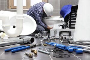 A plumber in a hard hat fixes a toilet in a tiled bathroom, surrounded by tools and plumbing parts.