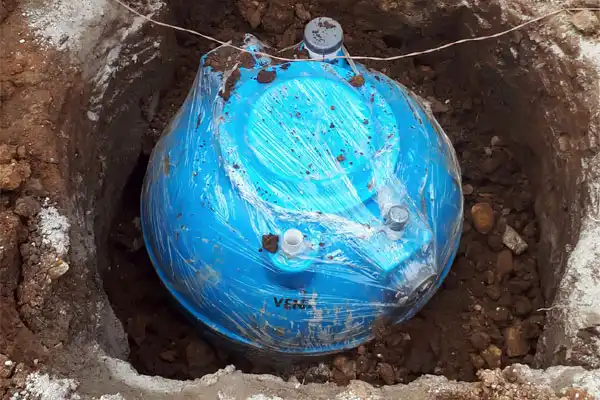 A blue underground water tank, reminiscent of a meticulous septic tank installation, is partially buried and shrouded in plastic wrap, surrounded by layers of soil and stones.