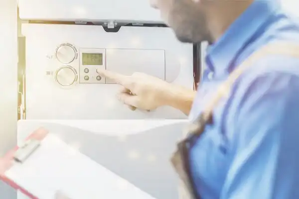 The technician in a blue shirt adjusts the settings on a boiler, holding a clipboard, as he prepares for a tankless water heater installation.