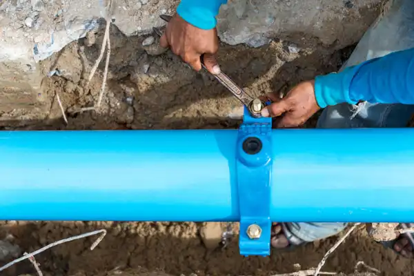 A person wearing a blue shirt tightens a clamp on an underground water pipe using a wrench, focusing intently on the water line repair.
