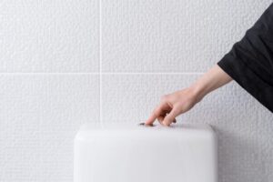 Hand pressing a chrome button on a white toilet cistern against a tiled wall.