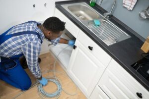 A plumber wearing a plaid shirt and blue overalls works under a kitchen sink, handling a coiled cable.
