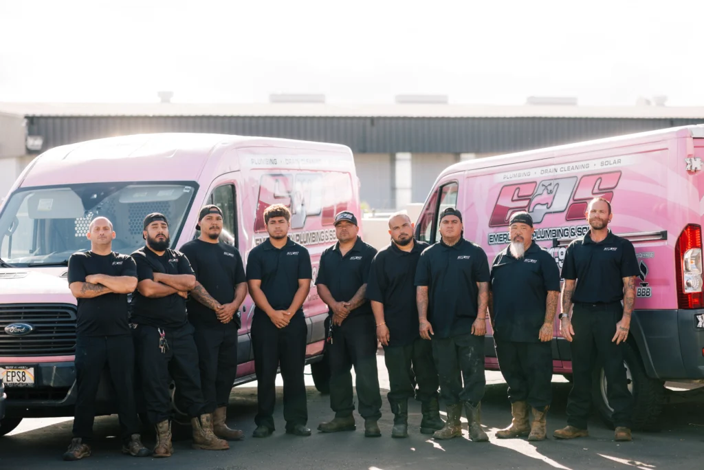 Nine workers in black uniforms stand in front of two pink plumbing company vans in a parking lot, ready to serve—learn more about us and our dedicated team.